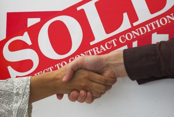 Two hands shaking in front of a sold property sign